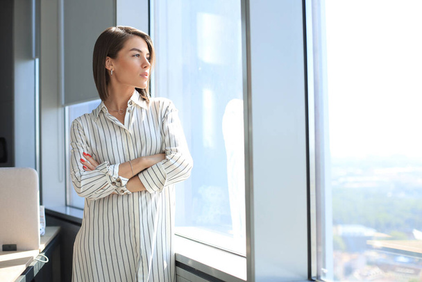 Attractive business woman in smart casual wear looking away and smiling while standing in the office. - Foto, afbeelding