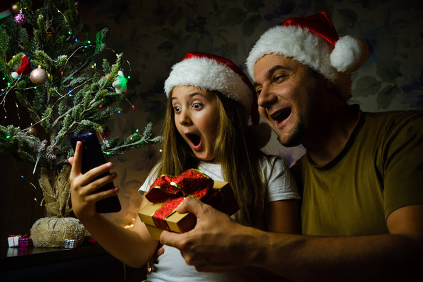 Enfant avec père chapeaux de Père Noël ayant un appel vidéo le jour de Noël, assis sur un canapé dans le salon avec arbre de Noël à la maison - Photo, image