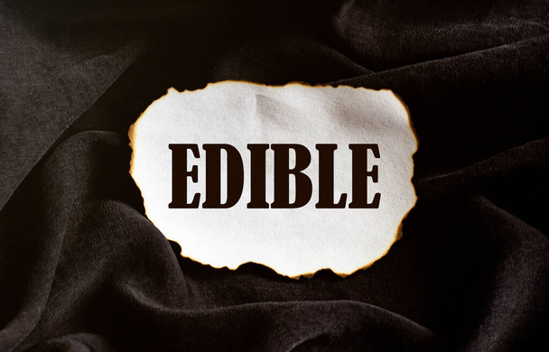 pedaço de papel branco queimado com texto EDIBLE em um fundo de tecido preto - Foto, Imagem