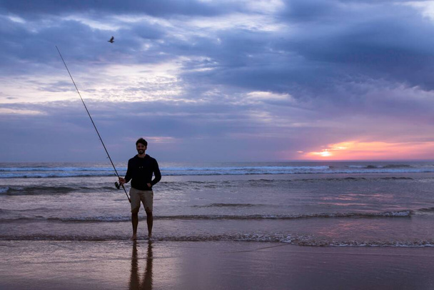Ein junger Mann fischt im offenen Meer entlang der Küste des Atlantiks an der Costa da Caparica in Lissabon, Portugal. Attraktiver Typ, der bei Sonnenuntergang am Strand die Angelrute in der Hand hält. Hobby-Lebensstil - Foto, Bild