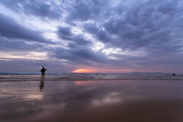 Een jonge man die vist in de open zee langs de Atlantische Oceaan aan de Costa da Caparica in Lissabon, Portugal. Aantrekkelijke kerel die de hengel vasthoudt bij zonsondergang op het strand. Hobby levensstijl - Foto, afbeelding