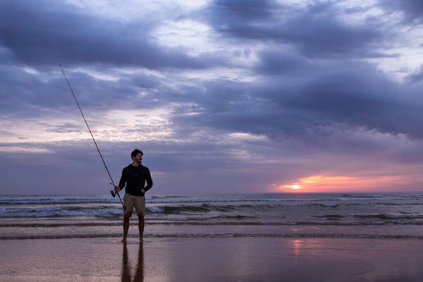 Un joven pescando en mar abierto a lo largo de la costa del Océano Atlántico en la Costa da Caparica en Lisboa, Portugal. Un tipo atractivo sosteniendo la caña de pescar al atardecer en la playa. Hobby estilo de vida - Foto, imagen