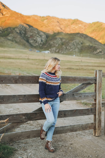 Молодая блондинка в нордическом свитере и джинсах возле старого деревянного забора на фоне гор - Фото, изображение