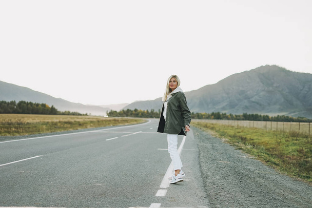 Schöne lächelnde blonde junge Frau Reisende in weißem Kapuzenpullover auf der Straße, Reise in die Berge, per Anhalter - Foto, Bild
