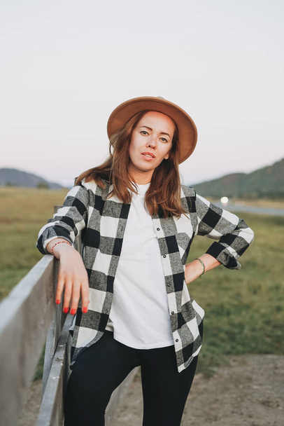 山の背景に古い木製の柵の近くに帽子と遊んだシャツを感じた若い女性 - 写真・画像