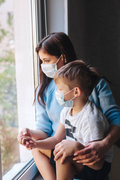 Mother and her son in protective masks very sad looking through window . Covid-19 pandemic and quarantine - Foto, Imagen