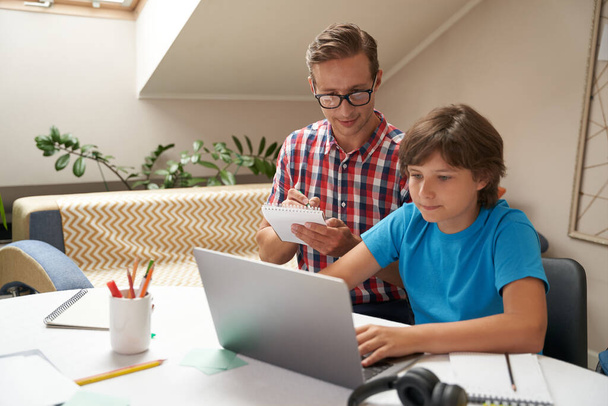 Otthon tanulási koncepció. Fiatal apa visel szemüveg és a kisfia néz video leckét online, férfi csinál néhány jegyzetek - Fotó, kép