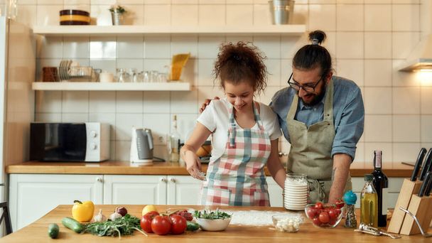 Young couple in love preparing the dough for making pizza with vegetables at home. Man and woman wearing apron, cooking together indoors. Hobby, lifestyle - Foto, immagini