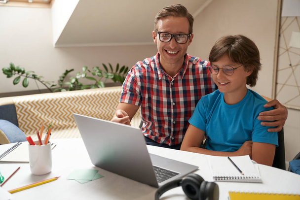 Junger glücklicher Vater hilft seinem Sohn bei den Hausaufgaben, sie benutzen Laptop und lächeln, Fernunterricht, Hausaufgabenkonzept - Foto, Bild