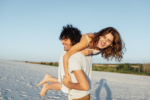 Beautiful couple having fun on the beach. Cheerful girl jumped on her boyfriends back - Zdjęcie, obraz