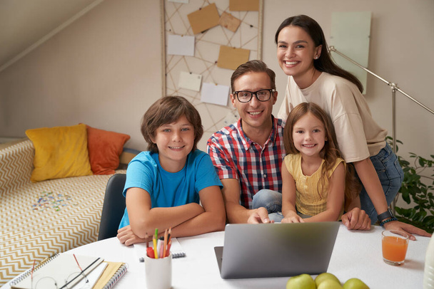 Портрет молодої щасливої сім'ї, яка дивиться на камеру і посміхається, роблячи домашнє завдання разом або навчаючись онлайн вдома під час ковадла 19 карантину
 - Фото, зображення