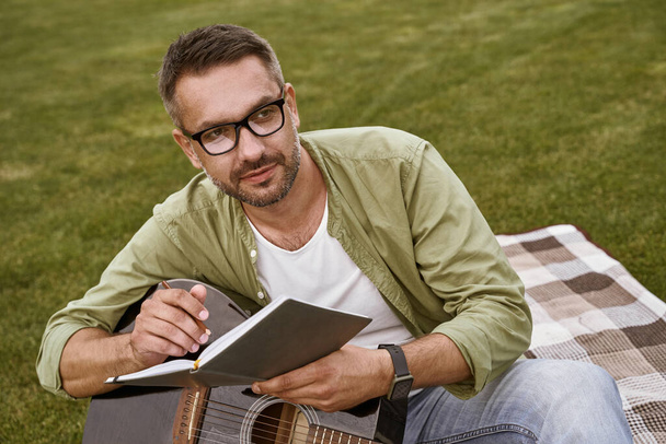 Male musician working in park. Young focused man wearing eyeglasses holding acoustic guitar and composing a song while sitting on a green grass outdoors - Photo, image
