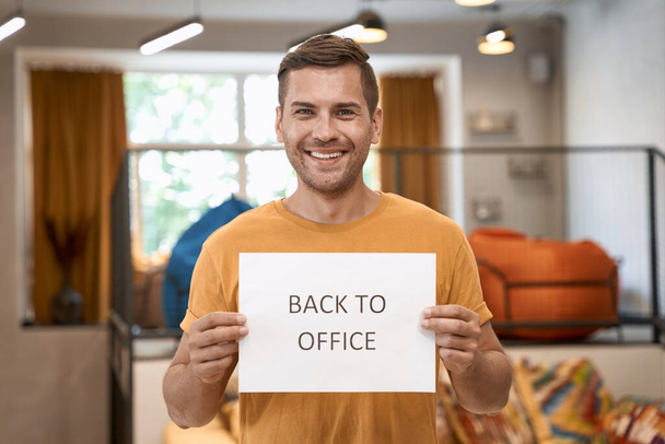 Reopening Business after lockdown. Young happy man showing paper with text BACK TO OFFICE at camera and smiling while standing in the modern coworking space - Zdjęcie, obraz