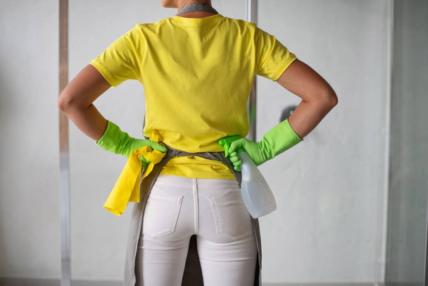 Pokojówka w fartuchu, żółte gumowe rękawiczki i żółta koszulka z rękami na biodrach. Trzyma butelkę sprayu i szmatę. Gotowy do drogi. Profesjonalne sprzątanie pomieszczeń i domów - Zdjęcie, obraz