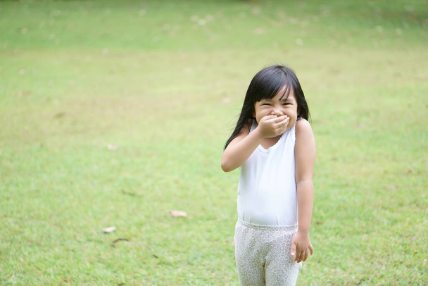 Азиатские дети мило или ребенок девушка стоять улыбка со смехом и руки закрыть рот для веселого удовольствия с пользоваться или кляп и секреты на зеленой траве луга пространство с носить белый жилет - Фото, изображение
