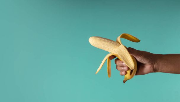 Banana Concept image. Mano sosteniendo una cáscara de plátano sobre el fondo azul, parece un arma. Foto de metáfora. Estilo limpio y mínimo - Foto, imagen