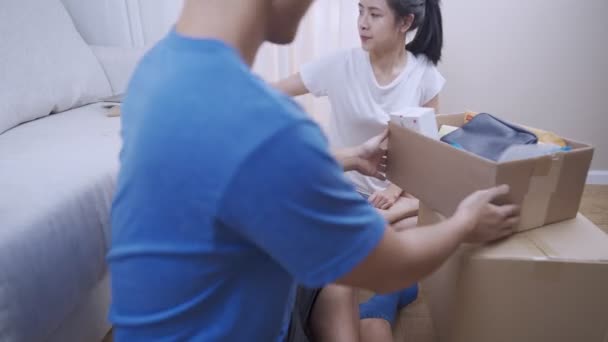 Young asian couple sorting things out of the parcel Carton box, sit down on floor moving inside new home living room, stack of cardboard box parcel container, Relocating moving into new place renting  - Footage, Video
