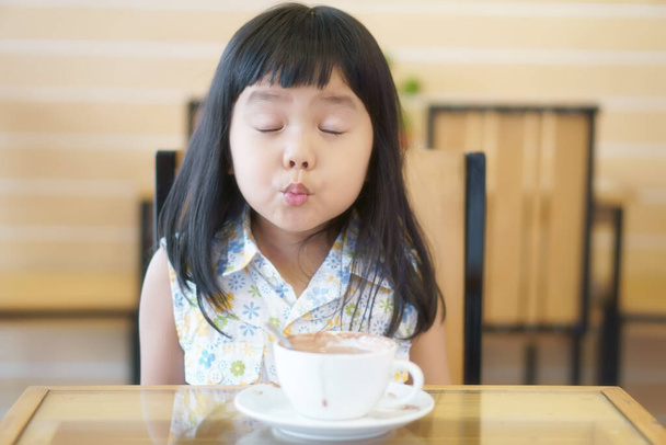 Bambini asiatici ragazza carina o bambino inviare baci con occhio vicino per amore e deliziosa cioccolata calda o bevanda in tazza bianca con divertimento felice per la colazione al mattino sul tavolo a casa o al ristorante caffè - Foto, immagini