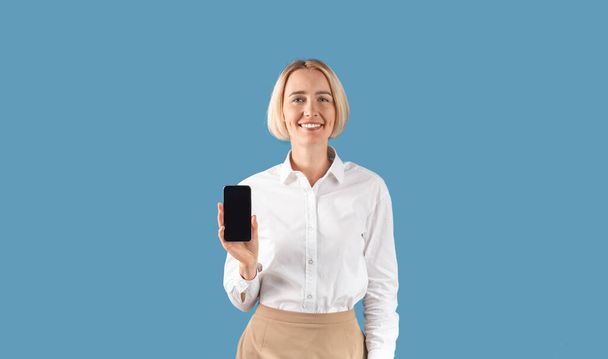 Sorridente amministratore delegato femminile in abbigliamento formale che mostra il telefono cellulare con schermo bianco su sfondo blu studio, panorama - Foto, immagini