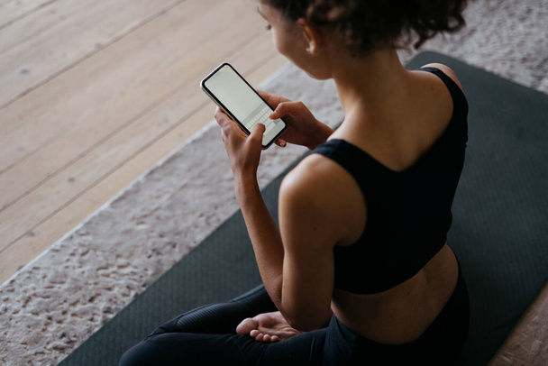 Magas látószögű kilátás felnőtt afro-amerikai nő, az online alkalmazás a modern okostelefon másolás helyet kijelző, ül a sport szőnyeg a nappaliban otthon, SMS-t és tartalmat blog - Fotó, kép