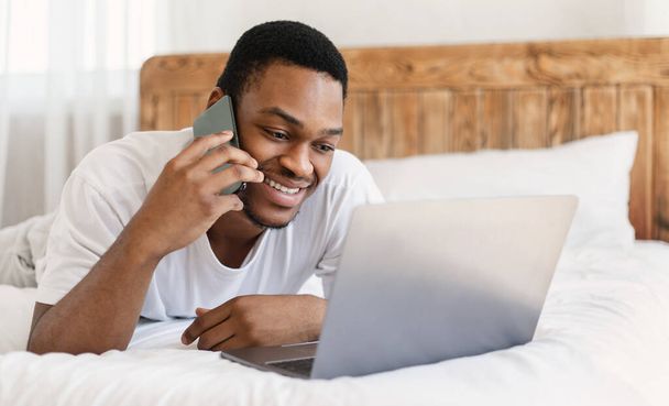 Hombre negro hablando en el teléfono celular usando el ordenador portátil acostado en la cama - Foto, Imagen