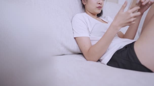 Aziatische jonge vrouw met behulp van smartphone leunend op de comfortabele bank ontspannen, online sociale media, mobiele applicatie, persoonlijke ruimte, benen gekruist op de knieën comfortabel op de bank, woonkamer - Video