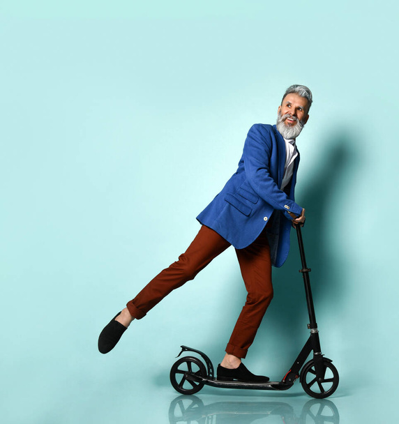 Gray-bearded aging man in white shirt, jacket, brown pants and loafers. Riding black scooter, smiling, posing on blue background - Photo, image