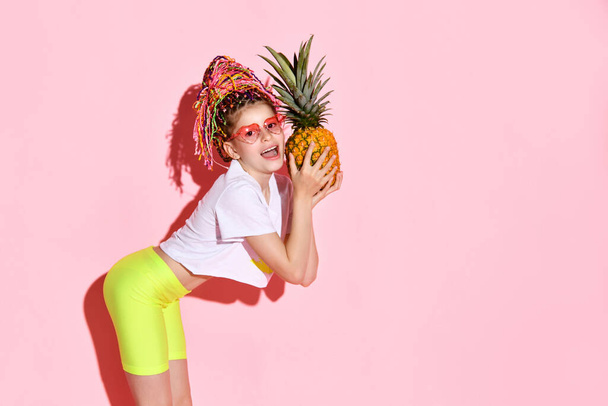 Funny schoolgirl in heart-shaped goggles with colorful afro bunches leaning forward with a pineapple in hands preparing to bite it, isolated on pink - Foto, Imagen