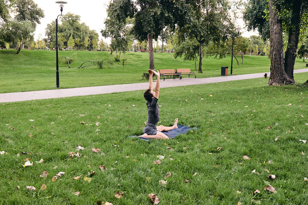 Young athletic man in sportswear doing yoga in the park. Practice asana outdoors. People exercising stretching on green grass with yoga mat. Fitness and healthy lifestyle. Concept of calm and meditation. - Zdjęcie, obraz