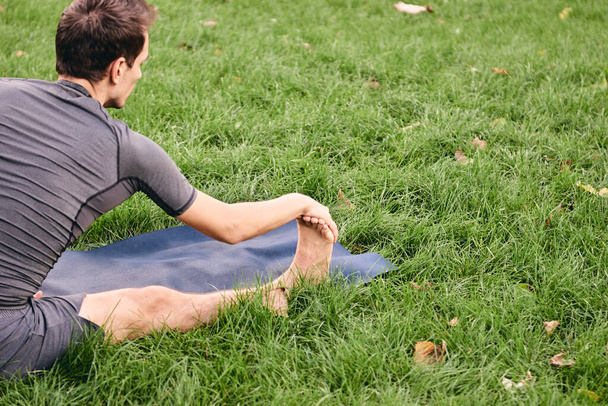 Młody sportowiec w odzieży sportowej uprawiający jogę w parku. Ćwicz asanę na świeżym powietrzu. Ludzie ćwiczący na zielonej trawie z matą do jogi. Zdrowy tryb życia. Koncepcja spokoju i medytacji. - Zdjęcie, obraz
