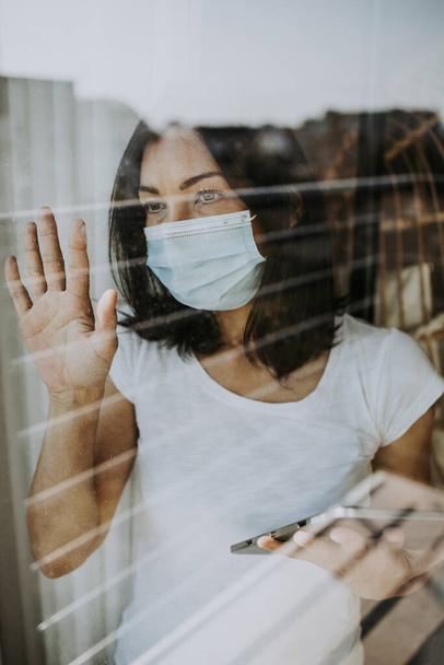 A frightened and worried woman looks out the window of her apartment or hospital room. She wears a protective mask on her face for fear of a dangerous and contagious virus. Quarantine and social distancing concept. - Foto, afbeelding