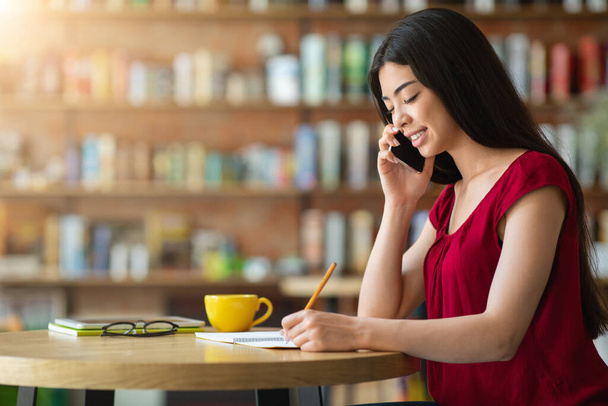 Усміхнена Азіатська Жінка говорить на мобільний телефон і приймає нотатки в кафе
 - Фото, зображення