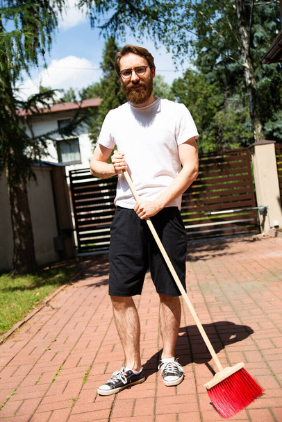 Sweeping outdoor. A young bearded man in glasses, cleaning the driveway in front of a property's entrance gate, with a broom, a sweeper. Cleanup works around the house on a sunny day. - Valokuva, kuva