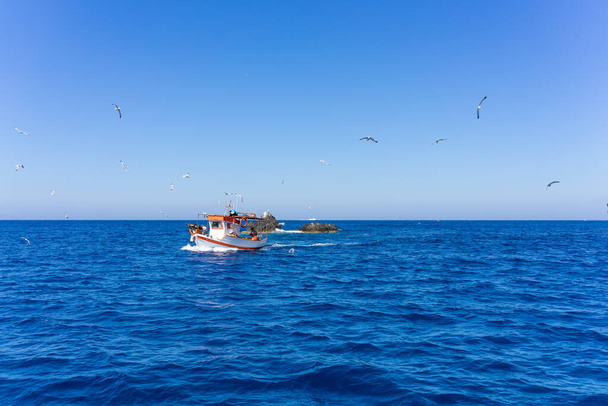Narancssárga és fehér halászhajó, utána sirályok az égei-tengeri tengeren. kék ég és napos nap - Fotó, kép