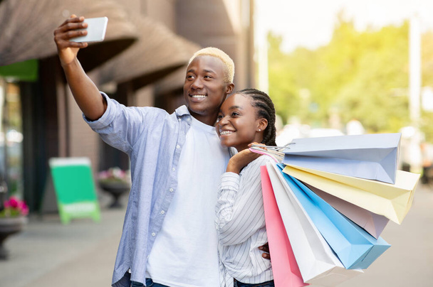 Foto de pareja feliz. Alegre afroamericano hombre hace selfie, novia con bolsas abrazos chico - Foto, imagen