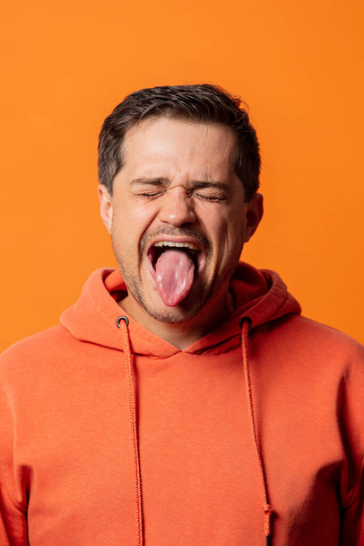 オレンジ色のパーカーの男はオレンジの背景に舌を見せ - 写真・画像