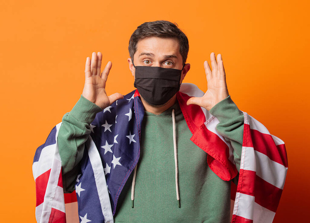 Yüz maskeli adam ve turuncu arka planda ABD bayrağı olan yeşil kapüşonlu. - Fotoğraf, Görsel
