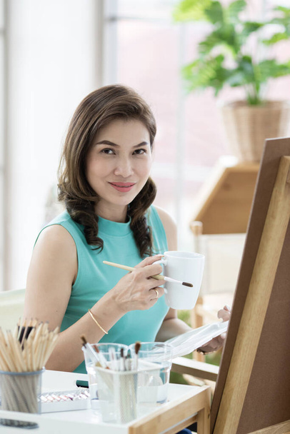 Belle dame asiatique femme assise et utiliser un pinceau pour l'image de la douleur dans la chambre. Idée de passe-temps, de détente ou de travail d'artiste de la maison. - Photo, image