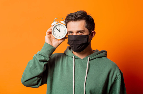 man in het gezicht masker en groene hoodie met wekker op oranje achtergrond - Foto, afbeelding