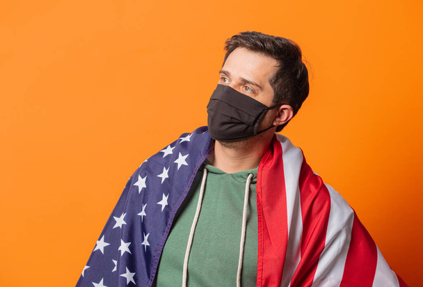 tipo en máscara facial y sudadera con capucha verde con bandera de EE.UU. sobre fondo naranja - Foto, Imagen