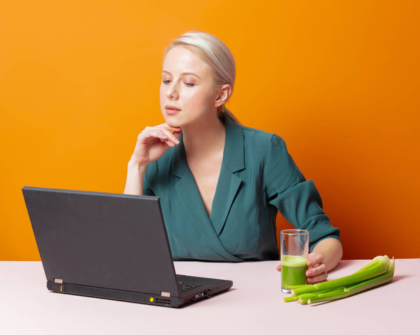 stylowa blondynka siedzi przy stole obok soku selerowego i laptopa  - Zdjęcie, obraz