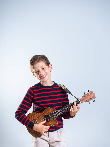 Jonge jongen speelt de ukelele, zijn voortanden ontbreken, geïsoleerd op grijze achtergrond, met kopieerruimte - Foto, afbeelding