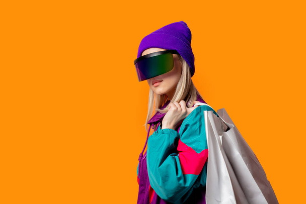 90 'ların eşofmanlı stil kadını ve turuncu arka planda alışveriş torbaları olan VR gözlükleri. - Fotoğraf, Görsel