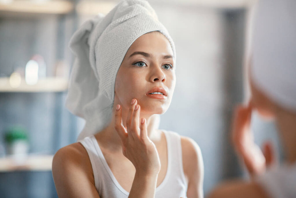 mooie jonge vrouw controleren haar huid voor rimpels in de buurt spiegel op badkamer - Foto, afbeelding