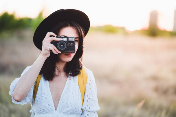 Young woman taking a photo with vintage film camera on the field. Hipster female with hat and white dress holding retro camera against sunset light from behind - Foto, afbeelding