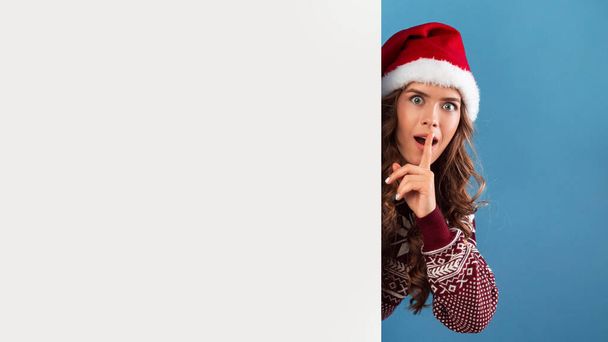 Dame im Weihnachtskostüm lugt hinter leerem Plakat hervor und macht stille Geste, blauer Hintergrund. Raum für Design - Foto, Bild