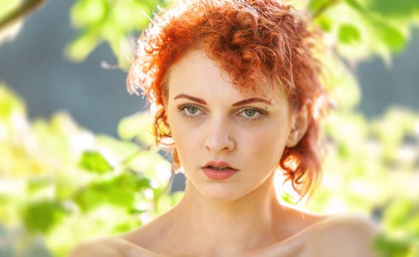 太陽の光に照らされた木々の葉の間に赤い髪の若い女性の肖像画 - 写真・画像