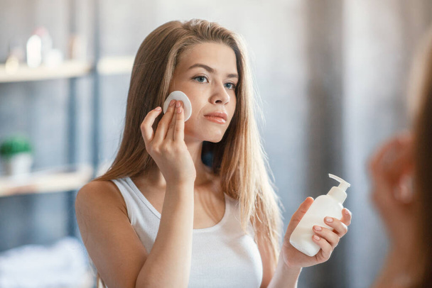 Hezká mladá žena pomocí obličeje tonikum nebo odstraňovač make-upu před zrcadlem v koupelně - Fotografie, Obrázek