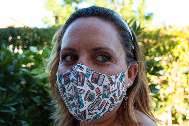 Vordergrundporträt einer attraktiven jungen Frau mit blauen Augen, die während des Coronavirus-Ausbruchs in einem Garten eine wiederverwendbare Maske trägt - Foto, Bild