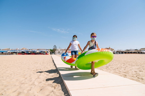 顔のマスクと眼鏡を持つ2人の女の子は、カラフルなインフレータブルマットレスホイールを腕の中でビーチへのパスを歩いてコロナウイルスパンデミック19の真っただ中で休暇を楽しむ - 写真・画像